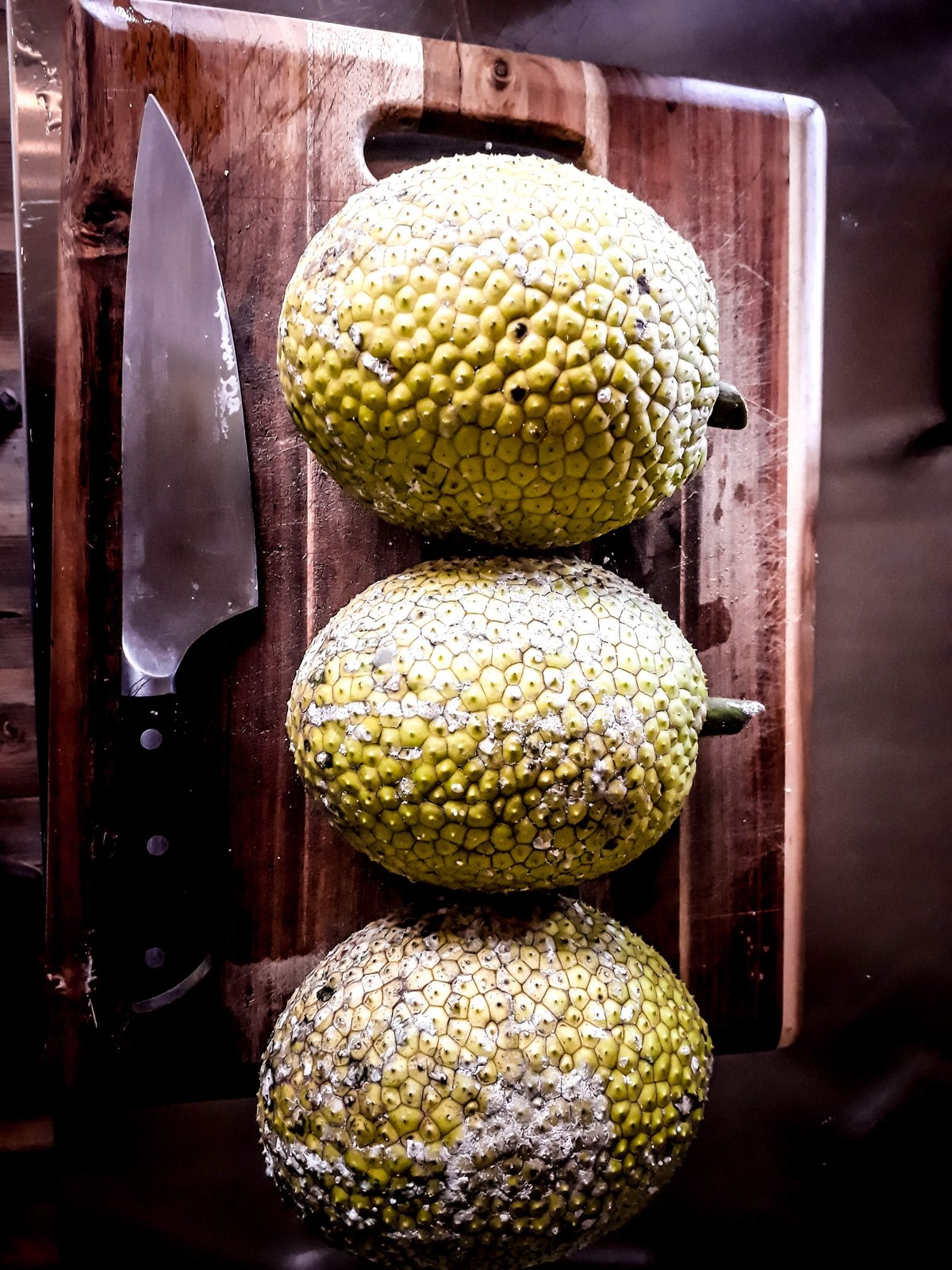 Three breadfruit stacked on a wooden board beside a chef's knife